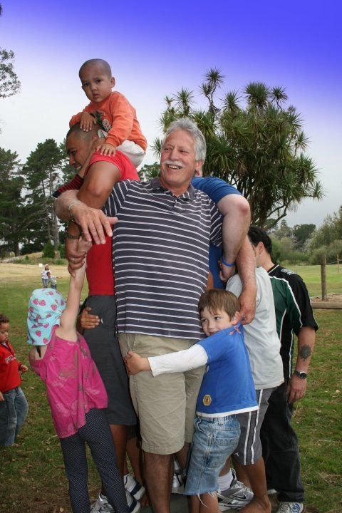 An older church member laughing as children climb on him and hug him at a park picnic.